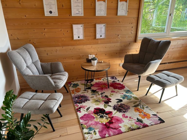 Praxisraum Heller Raum mit Holzwand, zwei Stühlen, einem Tisch und einem bunten Teppich.
