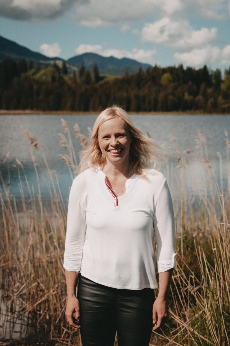 Franziska Howind Eine Frau mit blonden Haaren steht am Ufer eines Sees, im Hintergrund Berge und Bäume.